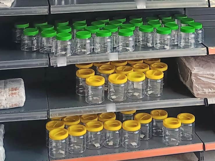 Rânduri de borcane transparente din plastic, cu capace verzi și galbene, pe rafturi
