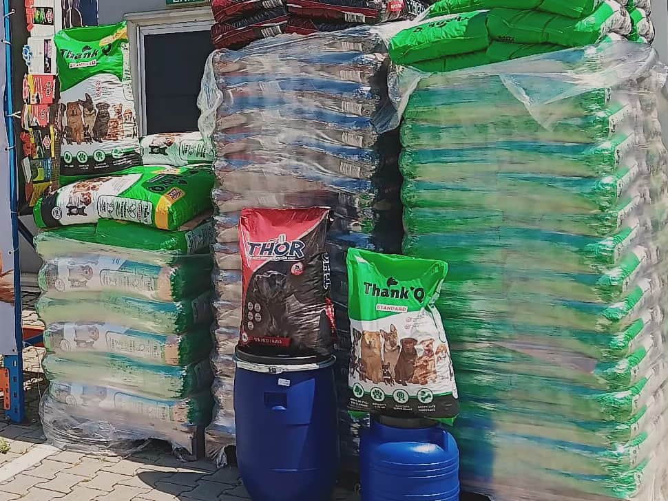 Paleți cu saci de hrană uscată pentru câini și pisici și două butoaie albastre, în fața depozitului