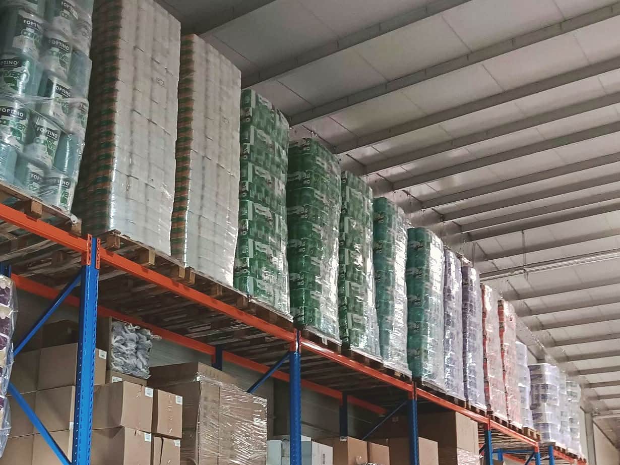 Culoar lung cu rafturi industriale portocalii și albastre, cu paleți de hârtie igienică și prosoape stivuiți până aproape de tavan