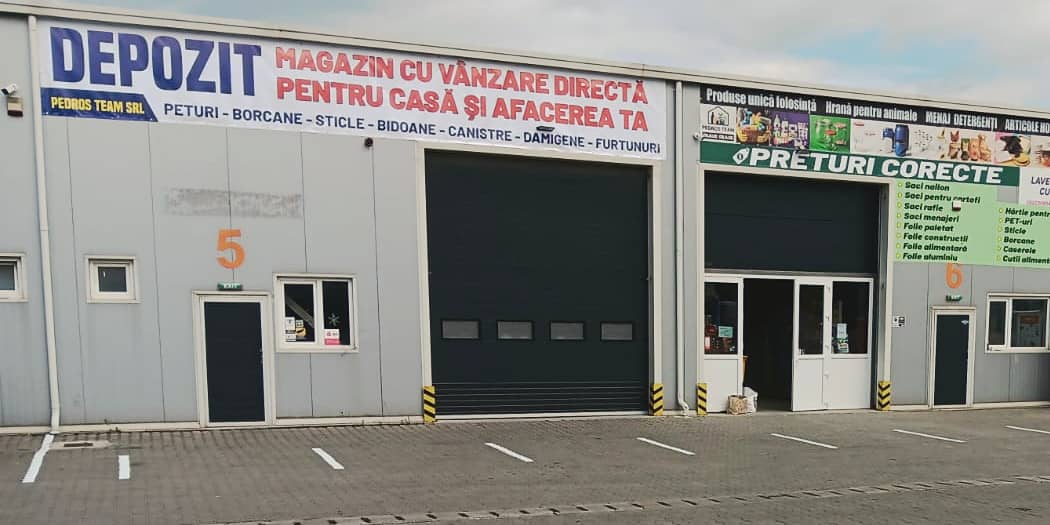 Fațada halei gri văzută din față: firma DEPOZIT, uși de garaj, ușa de sticlă deschisă și panoul verde „Prețuri corecte”